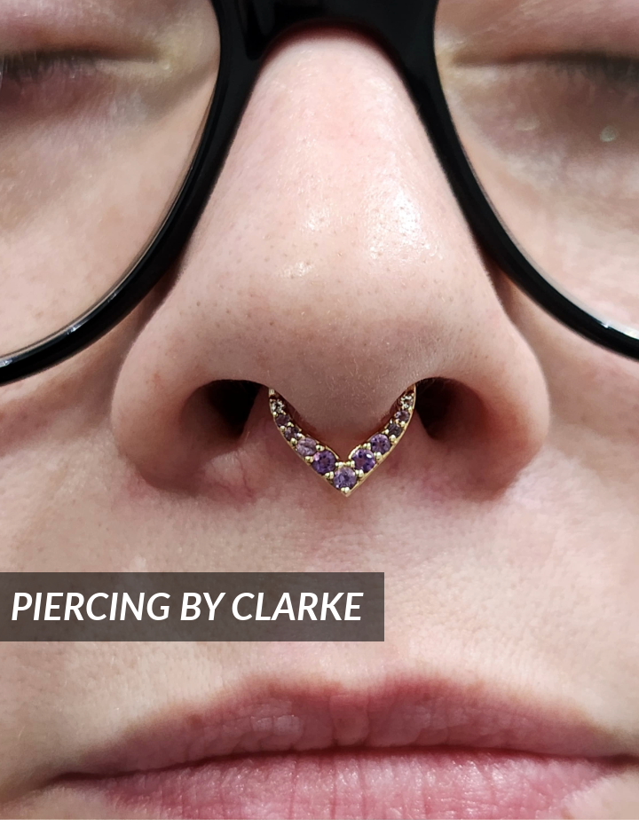 Close-up of a persons nose with a gold, V-shaped septum ring adorned with purple gems; the person is wearing black glasses. Text overlay reads PIERCING BY CLARKE.