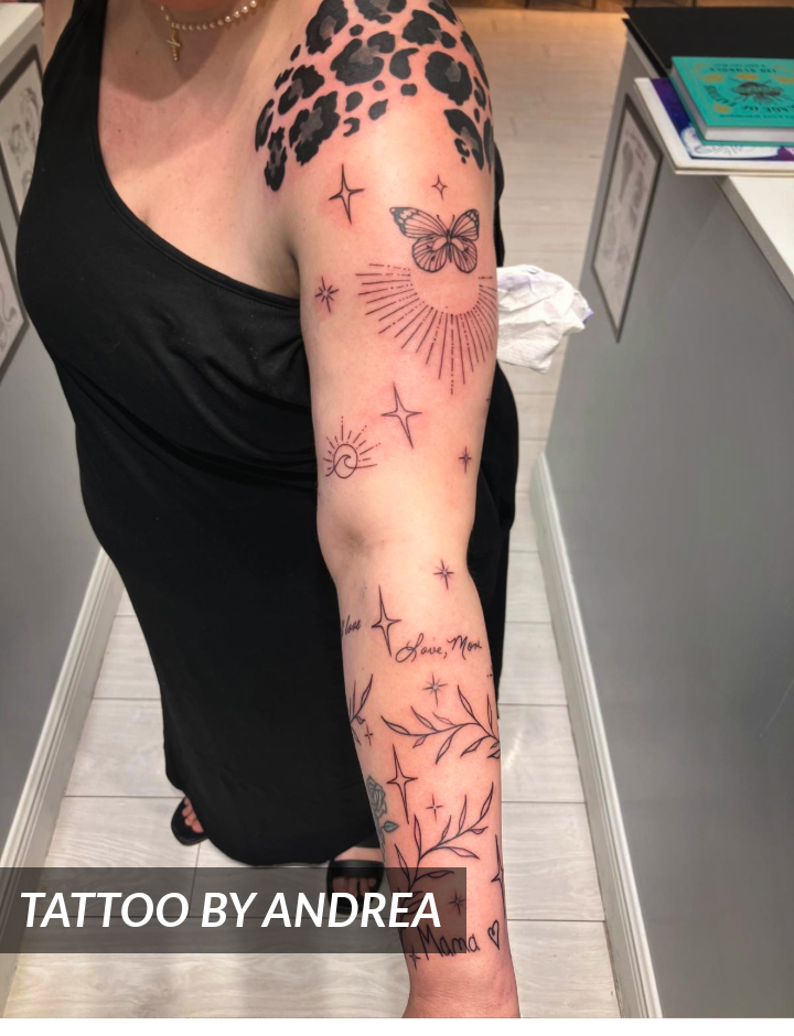 A person wearing a black dress displays a tattoo sleeve featuring a butterfly, stars, sun, leafy branches, script, and leopard spots on their shoulder. Text reads “TATTOO BY ANDREA.”.