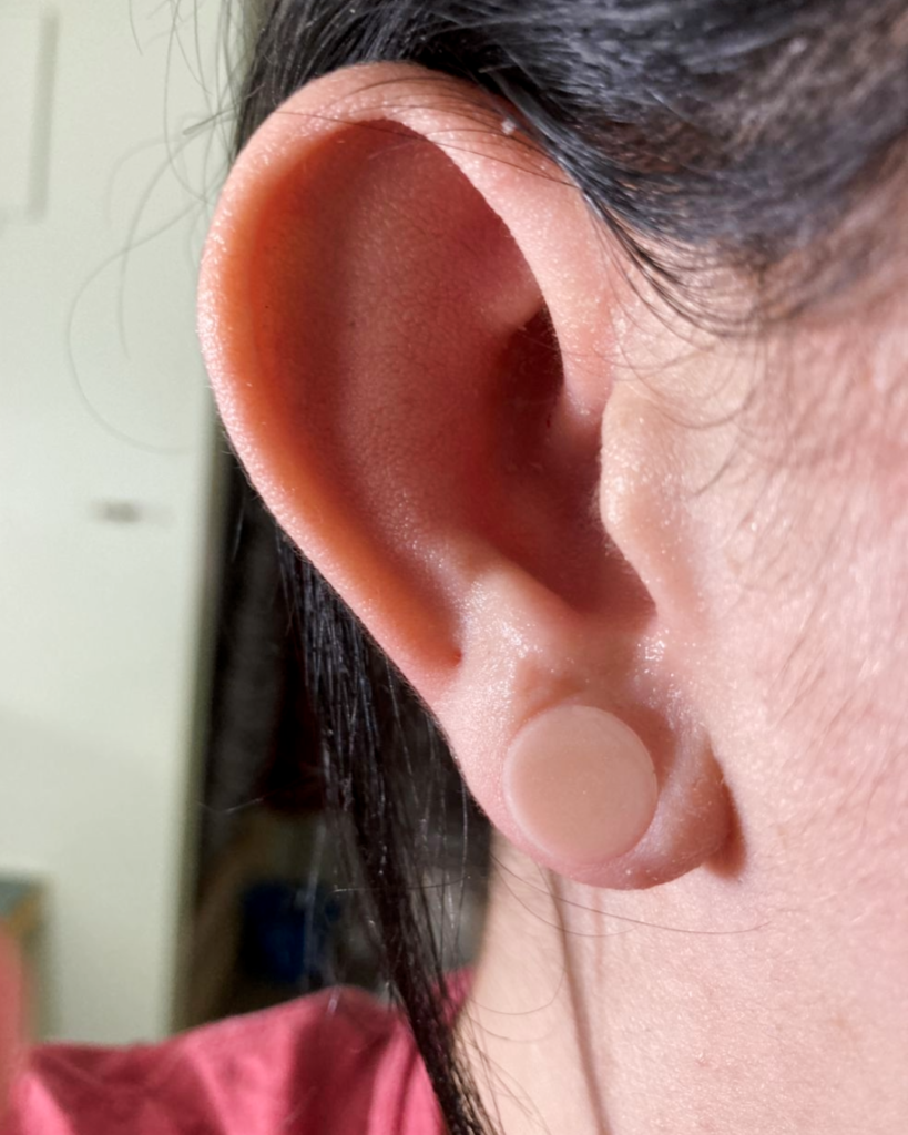 A close-up of a person's ear with dark hair, wearing a translucent, flesh-toned plug or gauge earring—featuring subtle lab grown diamonds—in a stretched earlobe piercing. The background is blurred.