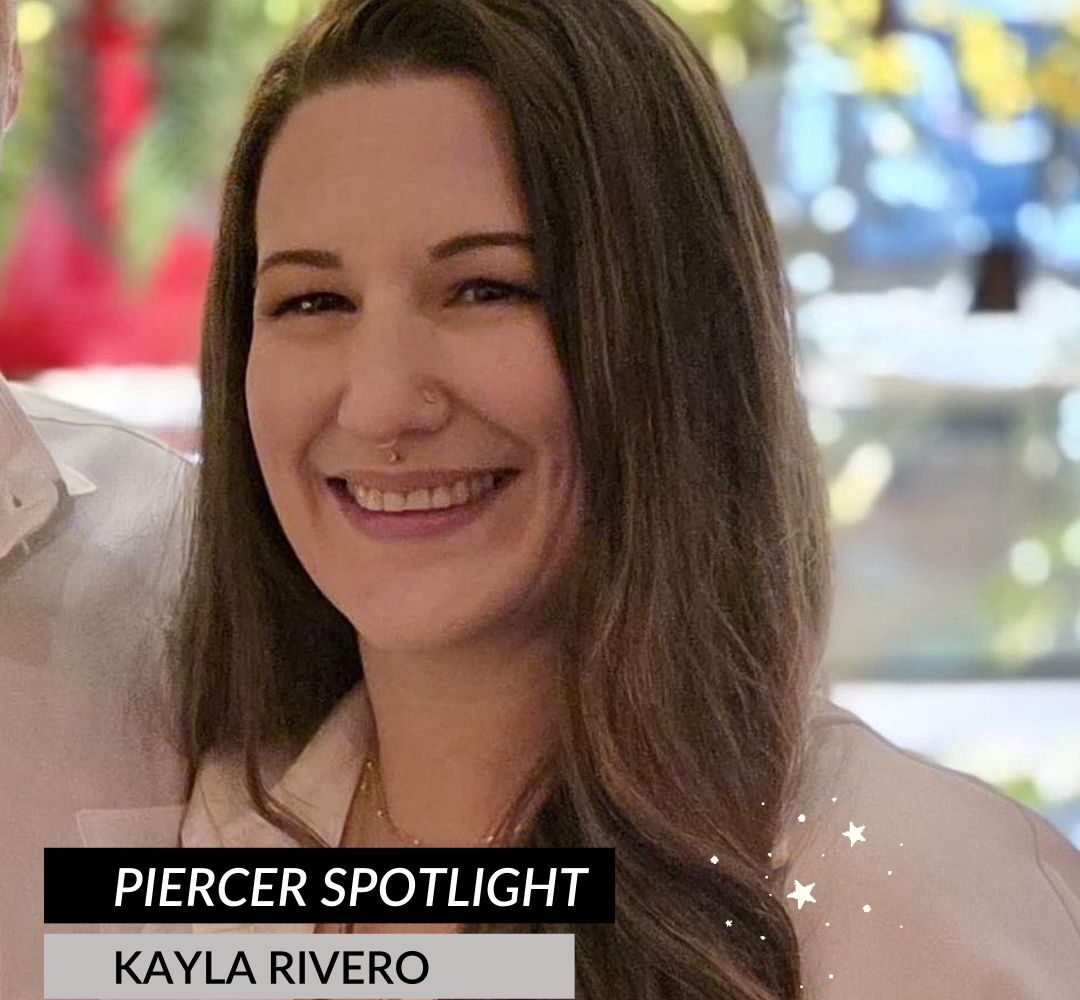 A woman with long brown hair smiles at the camera. She has a nose ring and is wearing a white top. Text on the image reads Piercer Spotlight: Meet Kayla Rivero, Guest Piercer. The background is softly blurred with colorful lights.