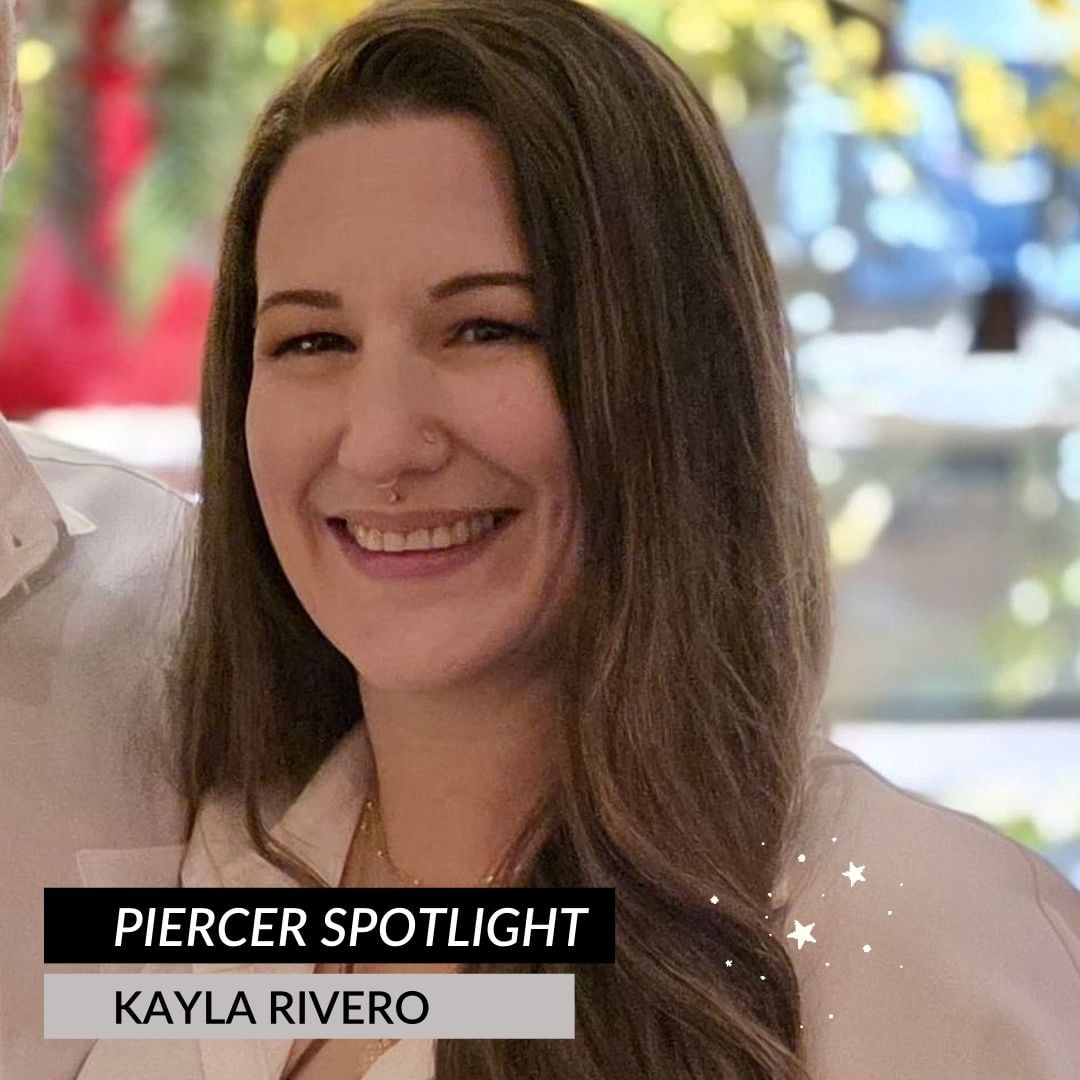 A woman with long brown hair smiles at the camera. She has a nose ring and is wearing a white top. Text on the image reads Piercer Spotlight: Meet Kayla Rivero, Guest Piercer. The background is softly blurred with colorful lights.