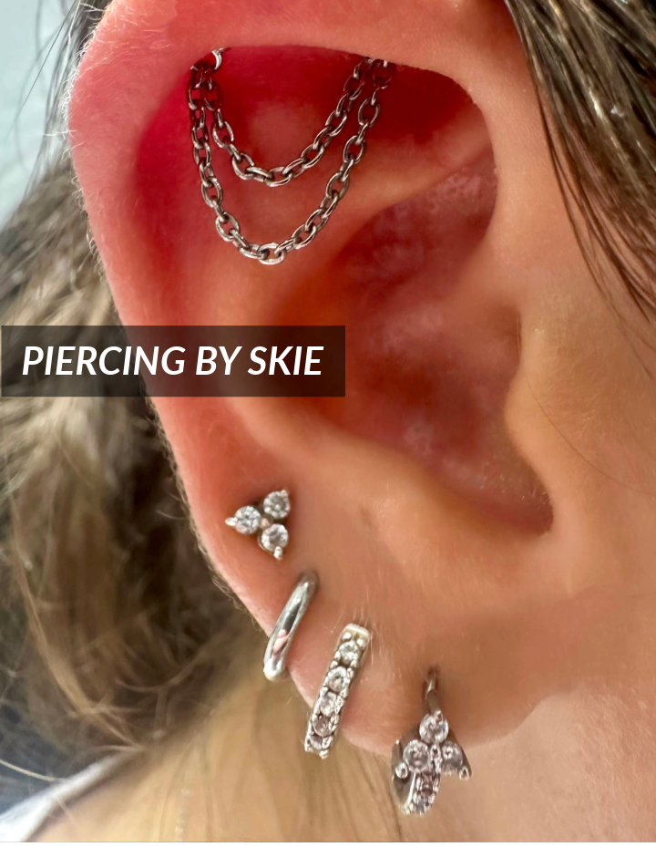 Close-up of an ear with multiple piercings, featuring a chain earring on the upper cartilage and various silver and diamond-studded studs and hoops. The text “PIERCING BY SKIE” appears over the image.