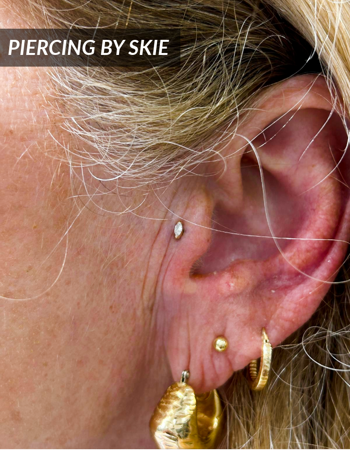 Close-up of a persons ear with three gold earrings and one small gemstone stud. The text PIERCING BY SKIE appears in the top left corner. The persons blonde hair frames the ear.