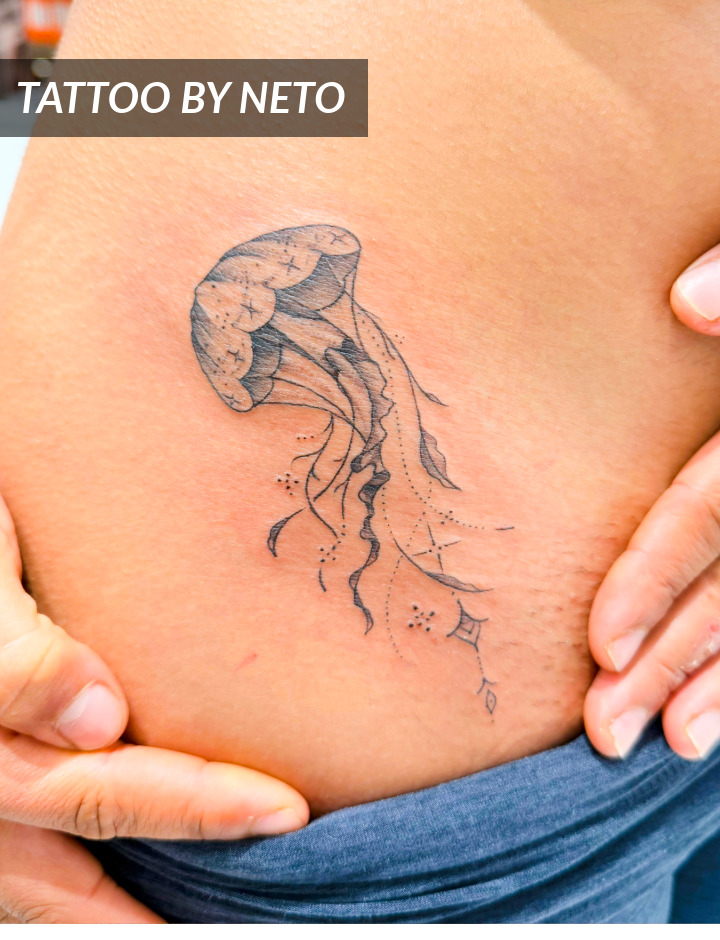 A close-up of a persons side shows a black ink jellyfish tattoo with delicate lines and dotwork. Hands gently pull garments aside to reveal the design. Text overlay reads, TATTOO BY NETO.