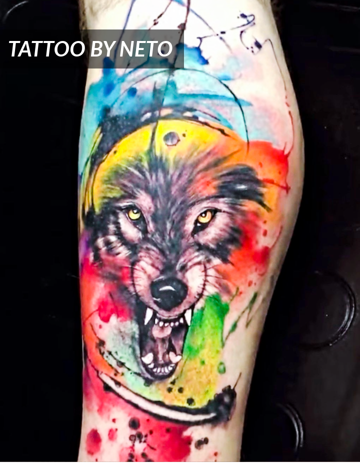 A colorful tattoo of a snarling wolf’s head, created in a watercolor style with splashes of vibrant blue, red, yellow, green, and black ink, on a person’s leg. Text reads TATTOO BY NETO.