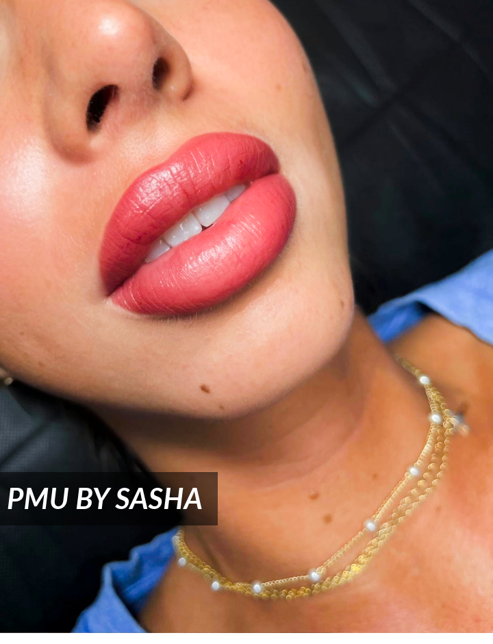 Close-up of a womans face showing full, well-defined lips with a bold pinkish-red color. She wears layered gold necklaces and a blue top. Text overlay reads PMU BY SASHA.