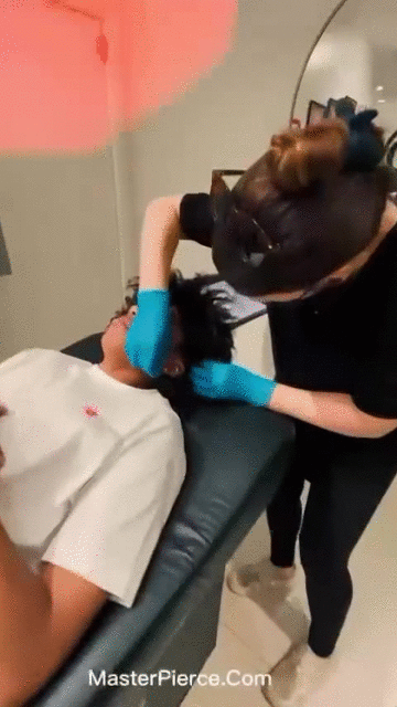 A person wearing blue gloves is piercing another persons ear while they lie on a padded bench in a clean, well-lit room. The website MasterPierce.com is visible at the bottom of the image.
