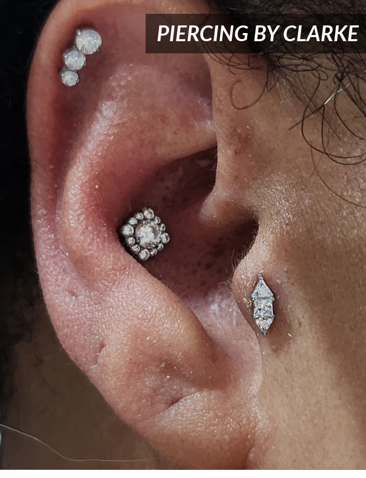 Close-up of an ear with multiple piercings, each adorned with sparkling stud earrings in various designs. Text overlay in the upper right corner reads PIERCING BY CLARKE.