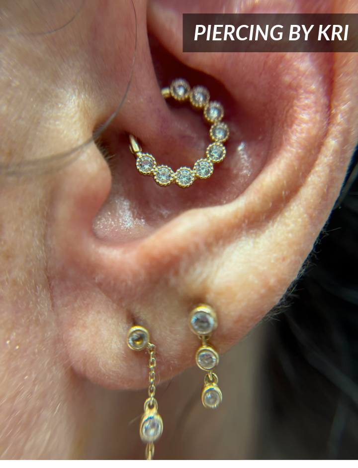 Close-up of an ear with a daith piercing featuring a gold and diamond hoop, and two dangling gold earrings with small clear gemstones. Text in the corner reads PIERCING BY KRI.