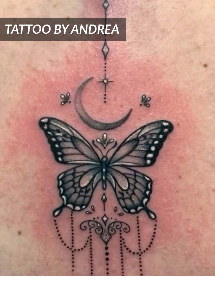 A black and grey tattoo on skin featuring a butterfly with ornate, geometric designs, a crescent moon above it, and decorative dots and lines hanging below.