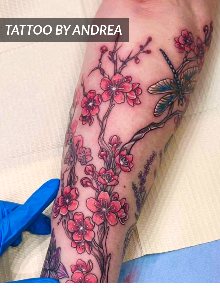 Close-up of a tattoo featuring pink cherry blossoms and a green dragonfly on a persons forearm. A gloved hand is visible next to the tattoo. The text TATTOO BY ANDREA appears in the top left corner.