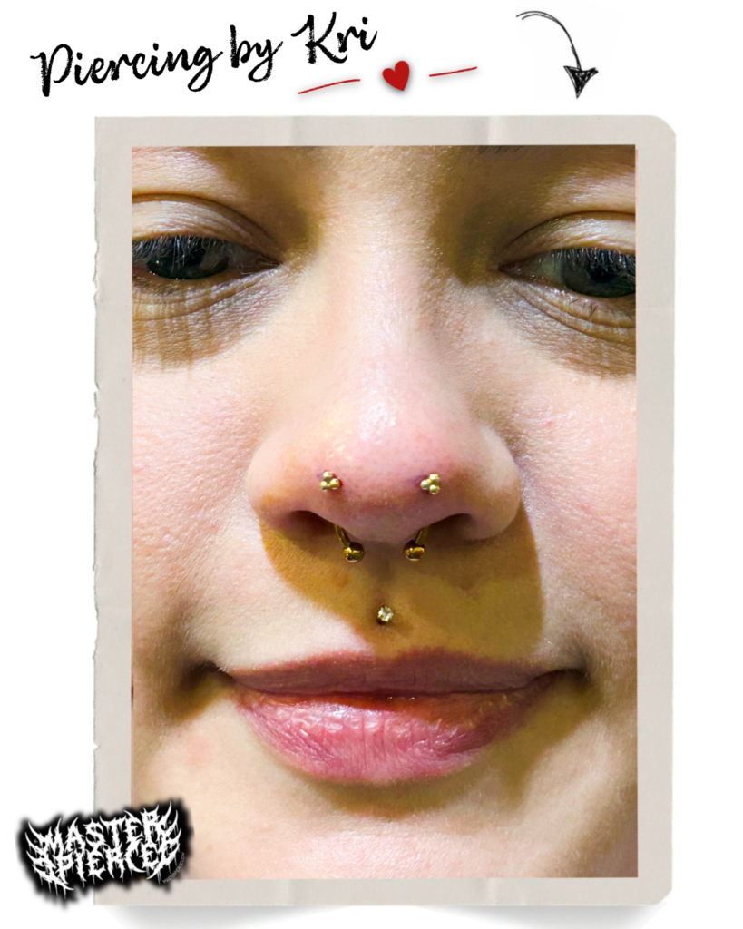 Close-up of a person’s face showing multiple gold-colored nose and septum piercings, with a small red mark on the nose. Decorative text above reads “Piercing by Kri” with a red heart and arrow.