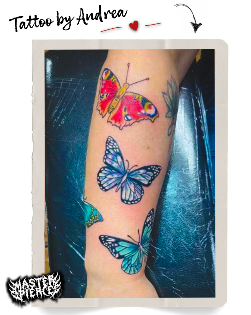 A forearm tattoo featuring three colorful butterflies—one red and yellow, and two in shades of blue and teal—on a persons skin. The image has a caption “Tattoo by Andrea” with decorative graphics.