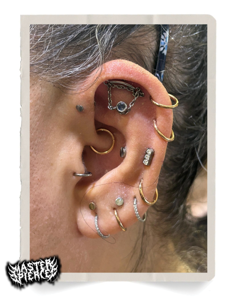 A close-up of an ear with multiple piercings, featuring gold and silver hoops, studs, and a decorative chain earring. The ear has various jewelry pieces in different shapes and textures, each expertly curated by Meet Kri Percing Artist to showcase a unique blend of style and craftsmanship.