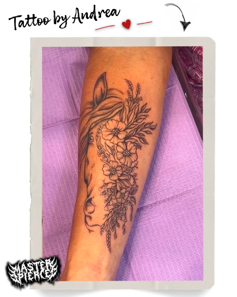 Forearm tattoo of a horses head blended with detailed flowers and leaves. The design is black ink and delicate, with Tattoo by Andrea written above the image on a paper-like border.