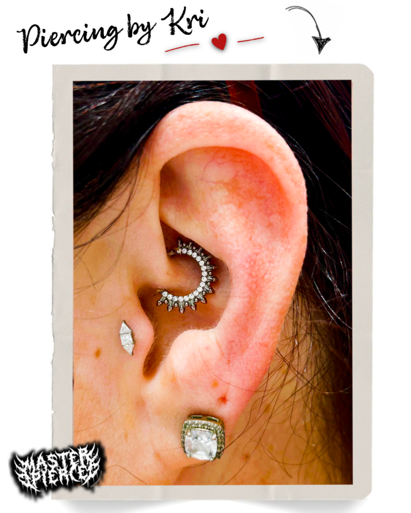 Close-up of an ear with three piercings: a decorative hoop in the conch, a diamond-shaped stud in the lobe, and a square gemstone stud below. Text says, Piercing by Kri with doodles and a “Wasted” graphic in the corner. Meet Kri Piercing Artist—where creativity meets precision.