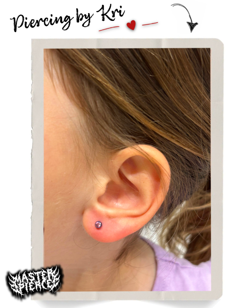 Close-up of a childs ear with a fresh lobe piercing and a pink stud earring. Text reads Piercing by Kri with a red heart, and a MASTERPIERCER logo appears in the bottom corner.