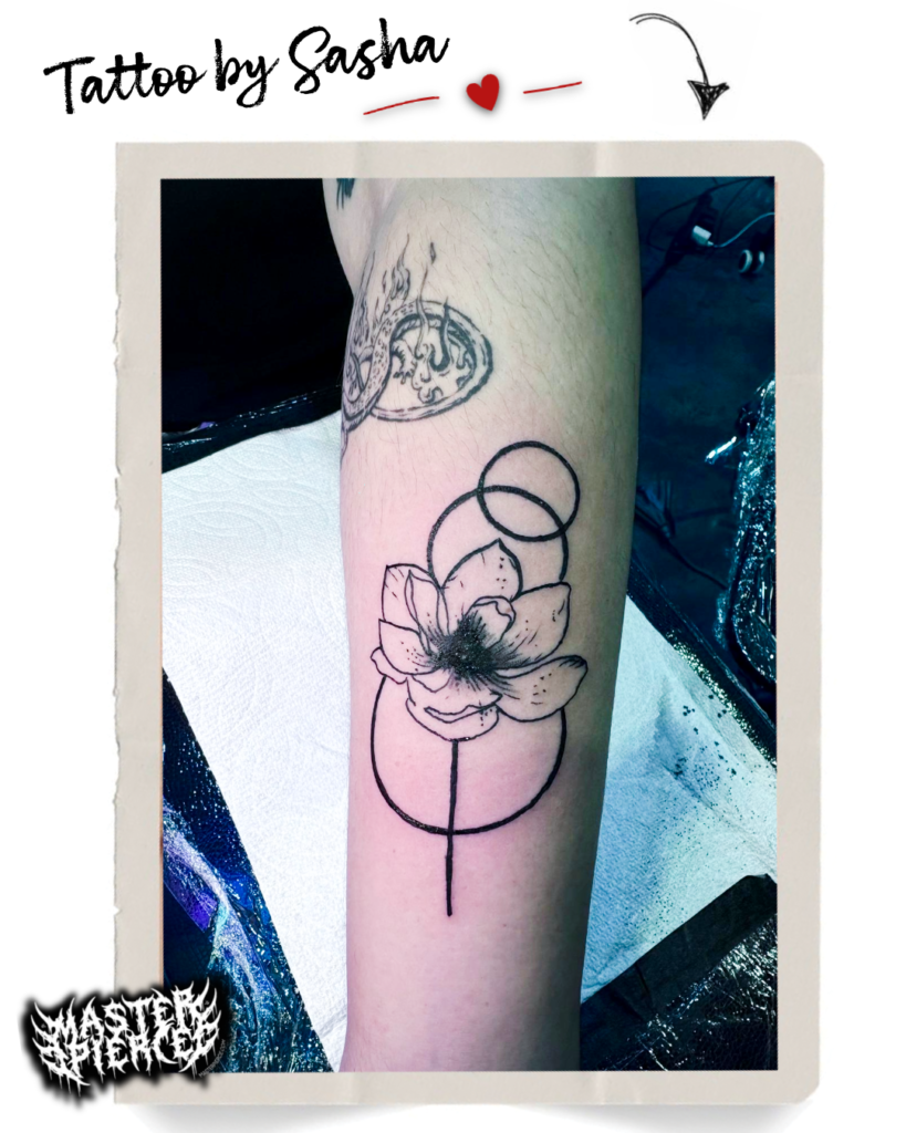 A forearm with a black ink tattoo of a large flower, abstract circular lines, and some fine shaded detail. Above it is an older tattoo featuring a round design. “Tattoo by Sasha” is written at the top of the image—meet Kri, piercing artist, whose creative influence shines through every piece.
