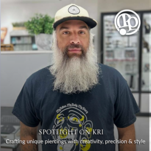 A bearded man in a hat and a graphic T-shirt stands in a piercing studio. Text reads: SPOTLIGHT ON KRI: Crafting unique piercings with creativity, precision & style. A logo is in the top right corner.