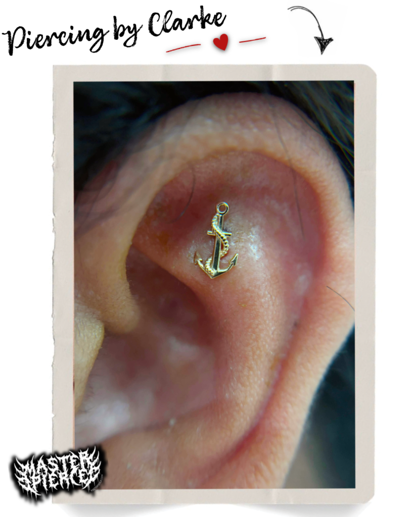 Close-up of an ear with a gold anchor-shaped piercing in the conch area. Decorative text reads Piercing by Clarke with an arrow and heart above the ear. A graffiti-style logo appears in the bottom left corner.