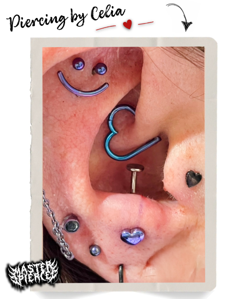 Close-up of an ear with multiple colorful piercings, including a blue heart-shaped hoop, a purple heart stud, a smiley face, and other metal jewelry. Piercing by Celia text appears at the top.