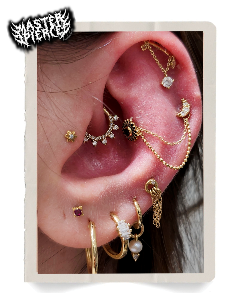A close-up of an ear with multiple gold, pearl, and diamond piercings, including chains, hoops, and studs in various designs. The ear has an ornate and decorative appearance.