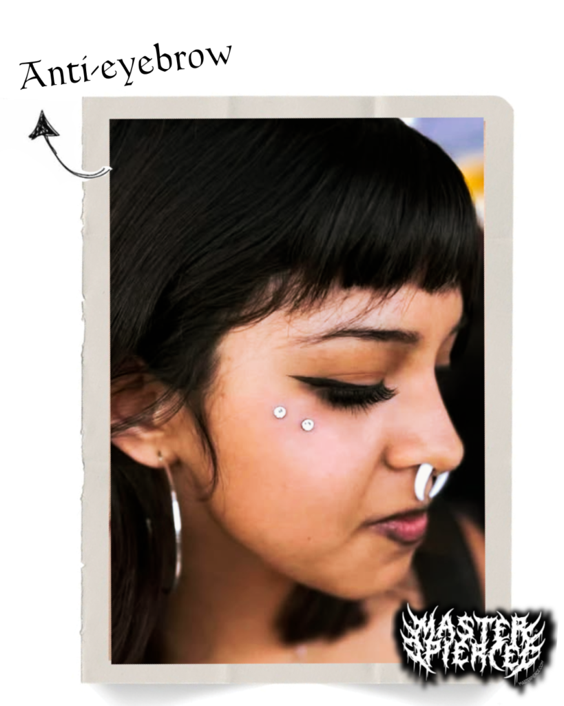 A person with straight black hair and bangs has two anti-eyebrow piercings below their right eye, a septum piercing, hoop earrings, and winged eyeliner. An arrow labels the anti-eyebrow piercings, offering a glimpse into the Ultimate Guide to Advanced Piercings.