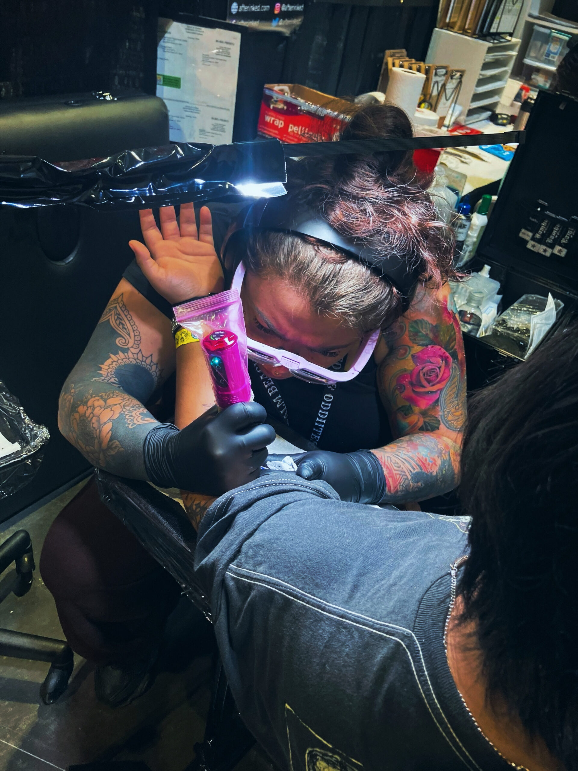 A tattoo artist with tattoos and gloves focuses on inking a clients arm at a busy studio, using a pink tattoo machine. The artist wears magnifying glasses and is surrounded by tattoo equipment.