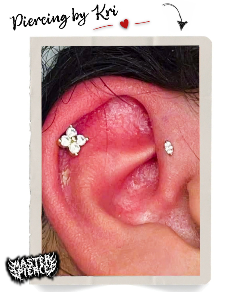 Close-up of a red ear with two fresh cartilage piercings: one with a flower-shaped cluster of gems, another with a single stud. Text reads Piercing by Kri with hearts and a decorative sticker in the corner.