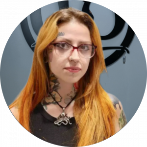 A person with long orange hair, glasses, and multiple tattoos, including on their neck and shoulders, stands in front of a gray wall with a black circular logo. They wear a black top and a necklace with an octopus pendant.