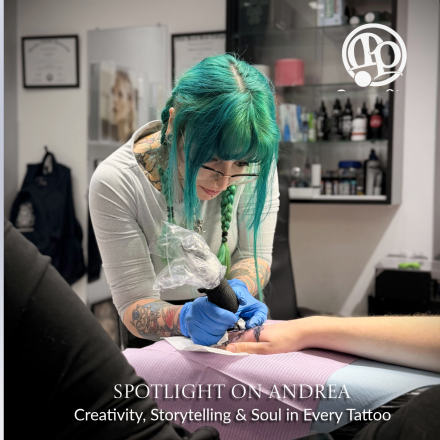 A tattoo artist with teal hair and glasses concentrates while tattooing a client’s arm in a studio. Text reads: “Spotlight on Andrea: Creativity, Storytelling & Soul in Every Tattoo.”.