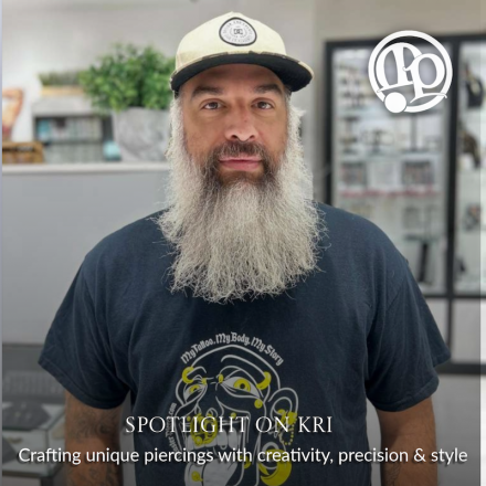 A bearded man in a hat and a graphic T-shirt stands in a piercing studio. Text reads: SPOTLIGHT ON KRI: Crafting unique piercings with creativity, precision & style. A logo is in the top right corner.