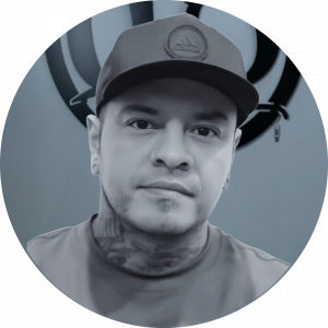 A man wearing a cap and a t-shirt looks at the camera. He has a tattoo on his neck and wears earrings. The background features a stylized black logo on a light blue wall.