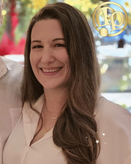 A woman with long brown hair smiles while wearing a white blouse and gold necklace. The background is softly blurred with warm colors, and a gold decorative logo is in the upper right corner.