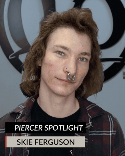 A person with light skin, long brown hair, and facial piercings, including a large septum piece, poses in front of a logo. Text at the bottom reads: Piercer Spotlight. Meet Skie Tattoo Artist: Skie Ferguson.
