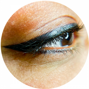 Close-up of a person's eye with black eyeliner and long eyelashes, highlighting detailed skin texture and eyebrow enhanced by Permanent Makeup (PMU). The image is cropped in a circular frame.