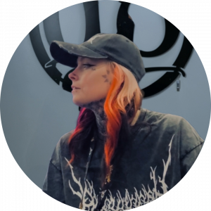 A person with long hair dyed pink and orange, wearing a black cap and a dark shirt with a white graphic, looks to the side. They have several face and neck tattoos. A large circular wall design is visible behind them.