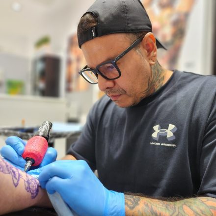 A tattoo artist wearing glasses, a black cap, and blue gloves is focused on tattooing a persons arm. He is holding a tattoo machine and working carefully on a purple stencil design.