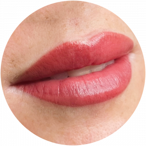 Close-up of a person's lips with smooth, reddish-pink Permanent Makeup (PMU) lipstick, slightly parted to show the edges of the front teeth. The light skin tone is framed in a circular crop.