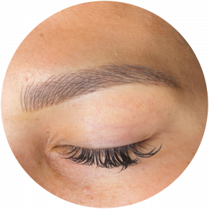 Close-up of a person's closed eye showing long curled eyelashes and a neatly shaped, filled-in eyebrow enhanced by Permanent Makeup (PMU) against smooth, light skin.
