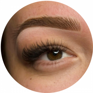 A close-up of a person’s brown eye with long, thick eyelashes and a well-defined, sculpted eyebrow enhanced by Permanent Makeup (PMU). The skin around the eye is clear and smooth.