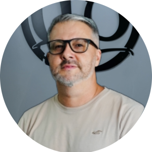 Meet Your Artist: A man with short gray hair and a beard, wearing black glasses and a light beige T-shirt, stands indoors in front of a gray wall with a circular black logo.
