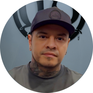 A man wearing a dark cap and shirt looks at the camera, tattoos visible on his neck. The gray background features a black circular design on the wall—Meet Your Artist in this striking portrait.