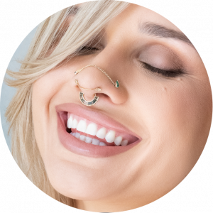 A smiling person with light skin and blonde hair wears unique nose jewelry connecting a nose ring to a septum ring, their eyes closed, teeth showing—showcasing creativity and style beyond just prices for piercing services.