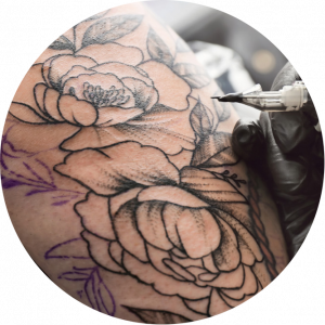 A close-up of a tattoo artist’s gloved hand using a tattoo machine to outline a floral design, with detailed petals and leaves. Prices for Piercing Services are available alongside custom tattoo options at the studio.