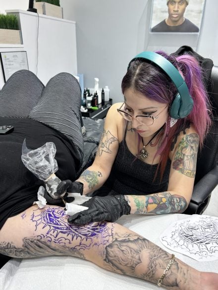 A tattoo artist with pink hair and headphones is focused on tattooing a client’s leg, following a purple stencil. The studio is clean and bright, with tattoo supplies and a portrait on the wall in the background.