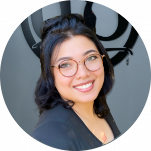 A person with medium-length dark hair, round glasses, and a nose ring smiles at the camera. They are wearing a black top and standing in front of a gray wall with a circular black design.